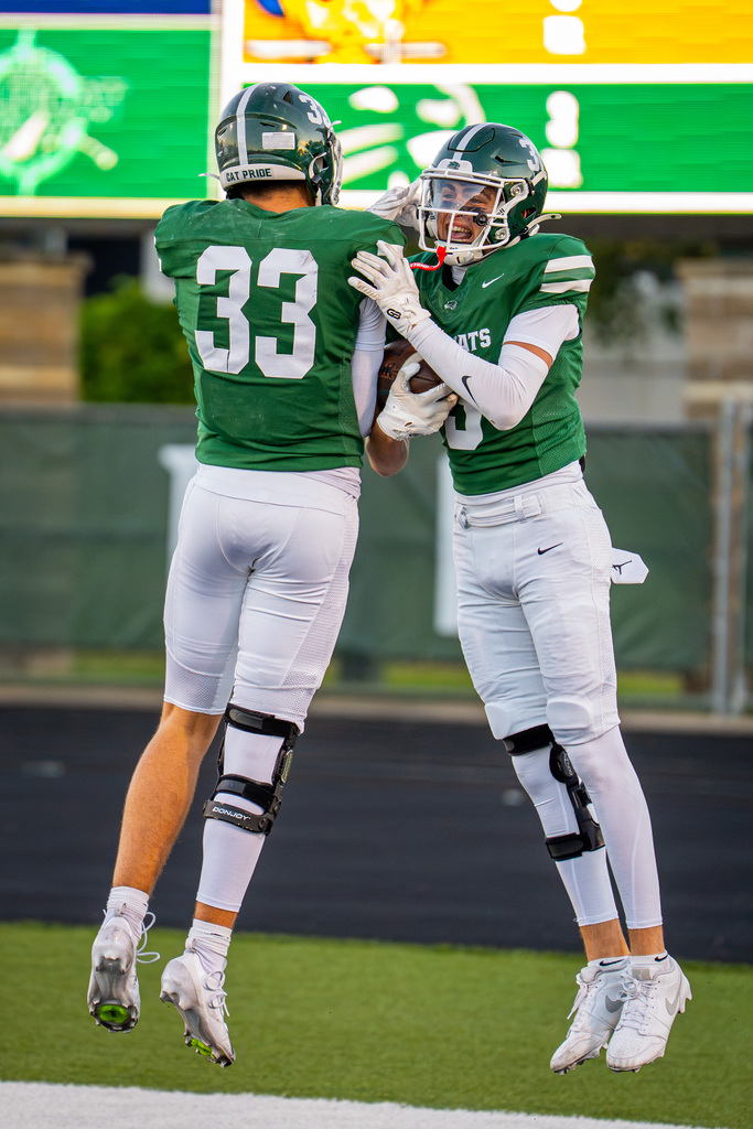 Two football players, wearing green and white uniforms, celebrate on a field with a green scoreboard in the background.