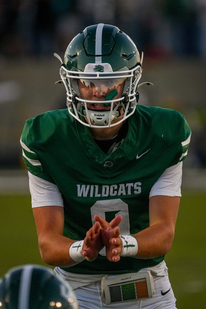Quarterback in a green Wildcats jersey and helmet is gesturing with his hands on a field.