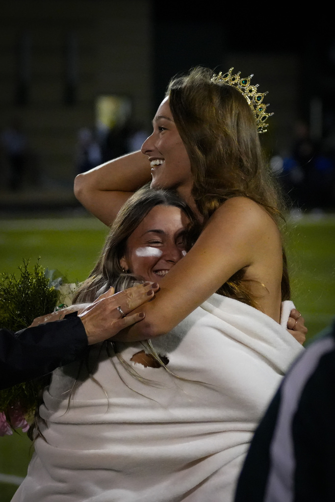 Woman with a gold crown smiles and hugs another woman with a white cloth. Background includes a stadium.