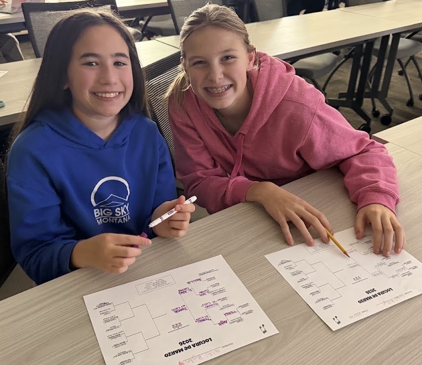 Two girls in hoodies sit at a table with papers and pens, smiling at the camera.