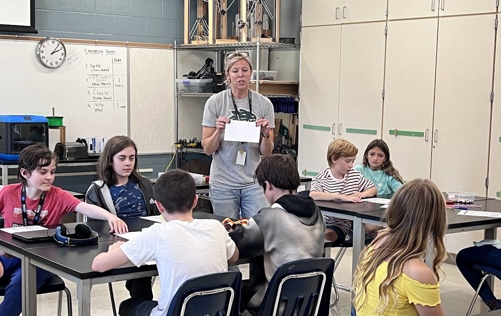 A classroom with children seated at tables. A teacher stands in front, holding a paper. A clock is on the wall.