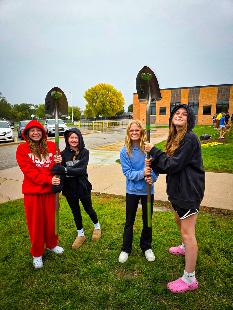 Four young women pose with shovels in a grassy area, with a school building and trees in the background.
