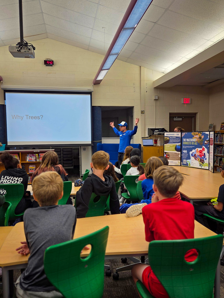 Classroom setting with projector screen at front, a man stands in front of it. Students sit in chairs at tables.