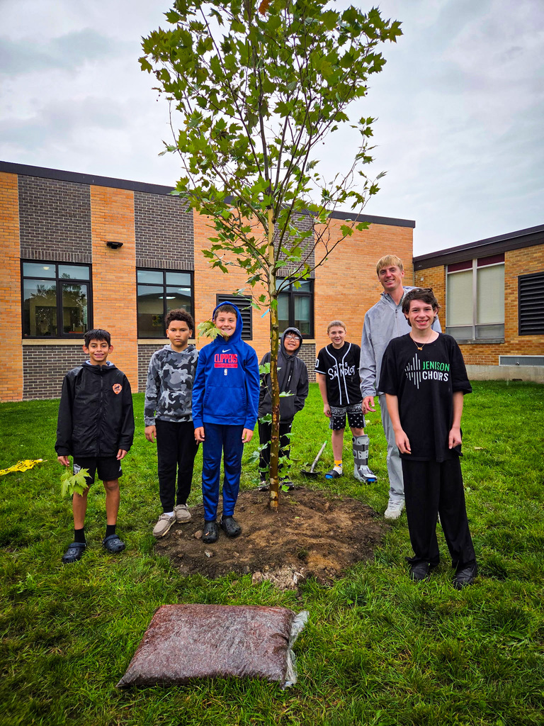 Seven children stand around a planted tree, smiling. A school building with brick walls and windows is in the background.
