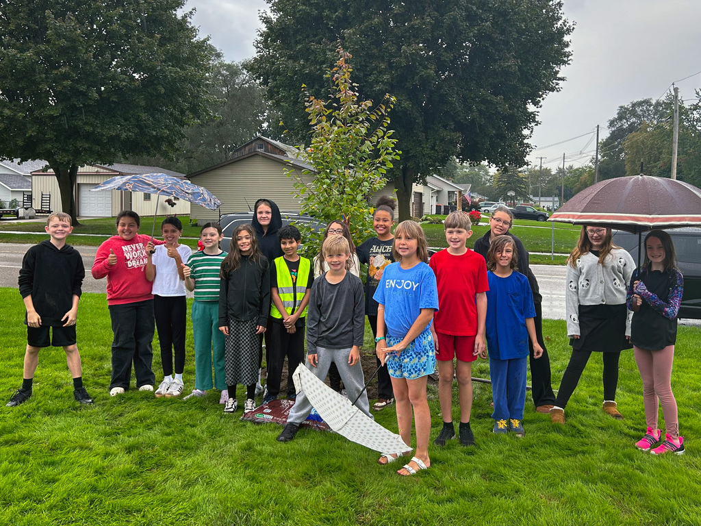 A group of young children standing outdoors on a lawn, some holding umbrellas, with trees and houses in the background.