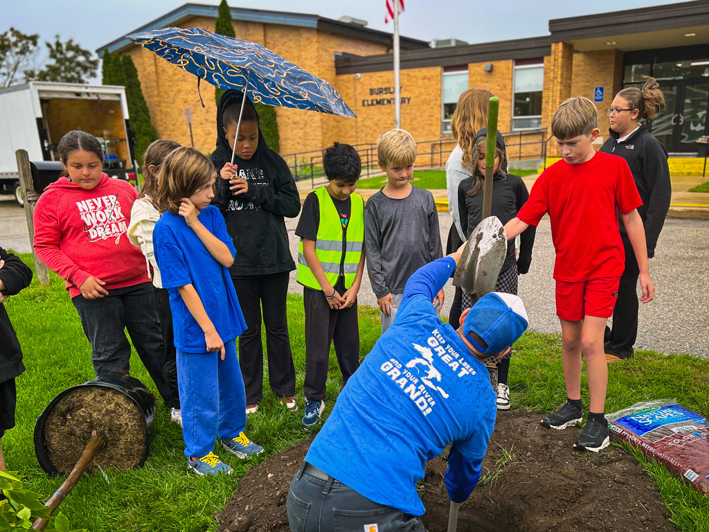 Kids in blue and red shirts watch a man plant a tree in a hole. Behind, a building is visible.
