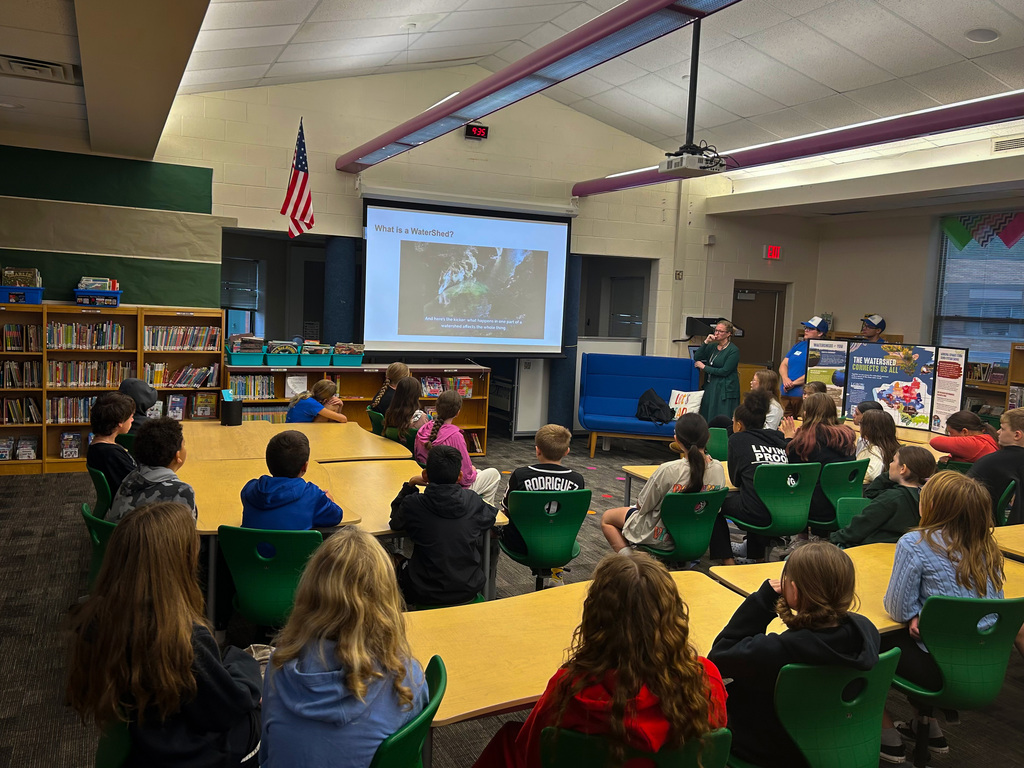 A classroom with students seated at tables watching a presentation on a projector screen. An adult stands by the screen.