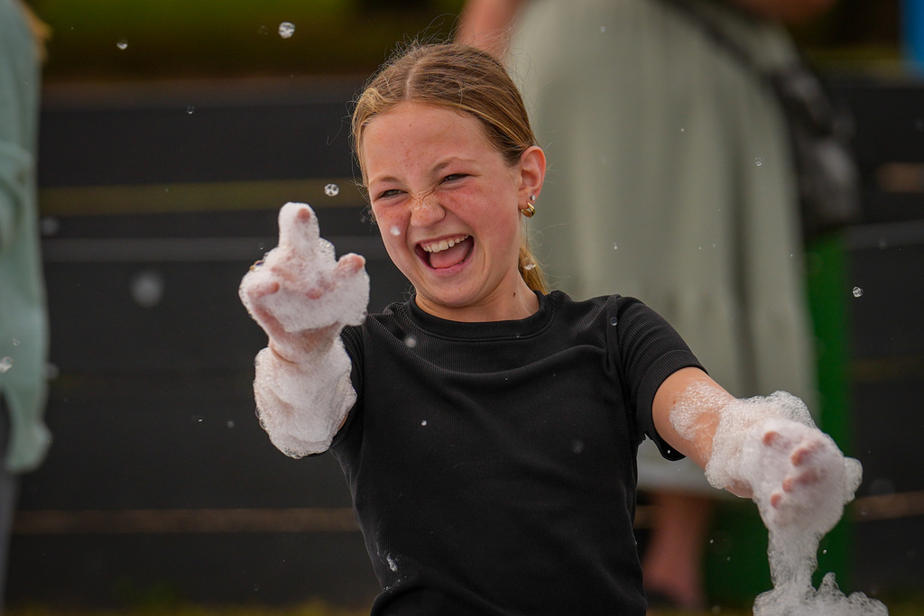 A girl in a black shirt holding foamy bubbles and smiling, with someone standing behind.