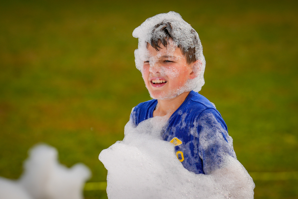 A young boy in a blue shirt with foam on his head, smiling and standing in a grassy area.