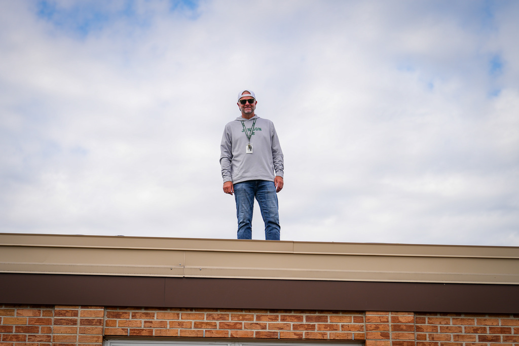 Principal in white shirt and sunglasses stands on rooftop of brick building under partly cloudy sky.