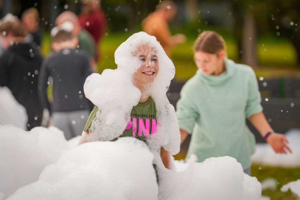 Children playing in a cloud of bubbles at an outdoor event, with people standing and walking in the background.