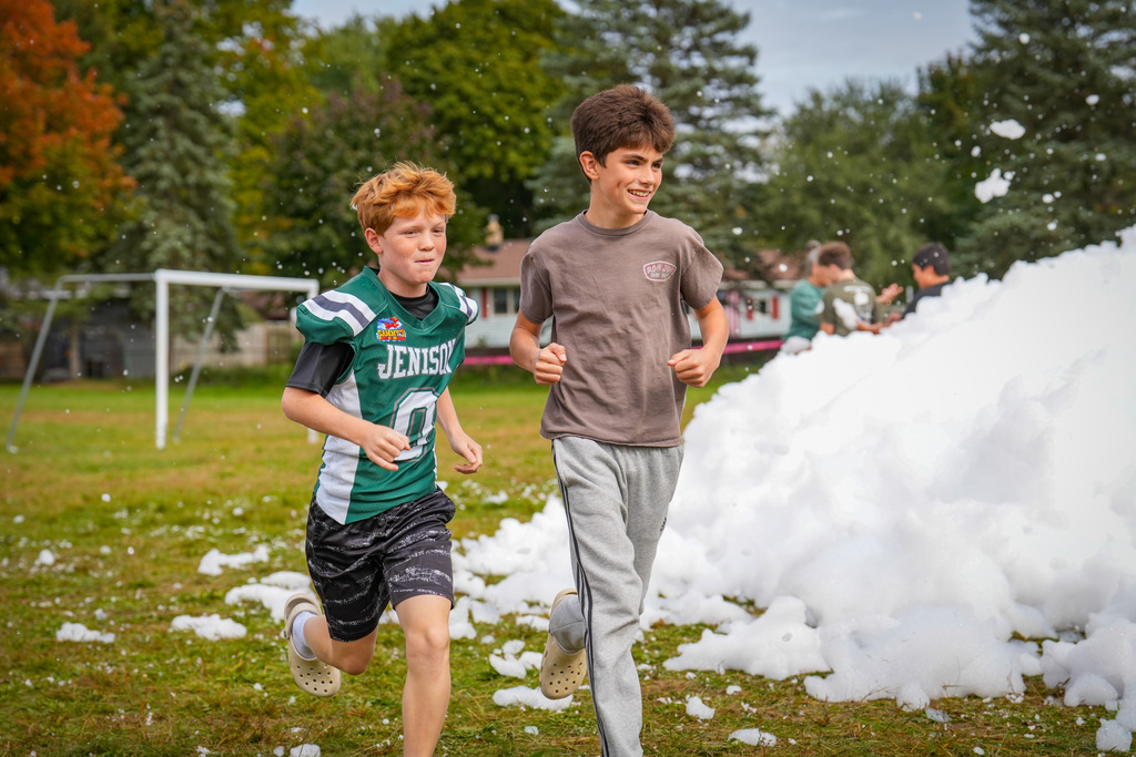 Two children running through a cloud of foam on a field with a soccer goal and trees.