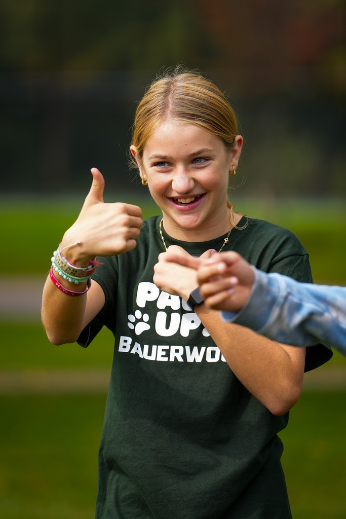 A young girl with blonde hair smiles, giving a thumbs up while wearing a green shirt.