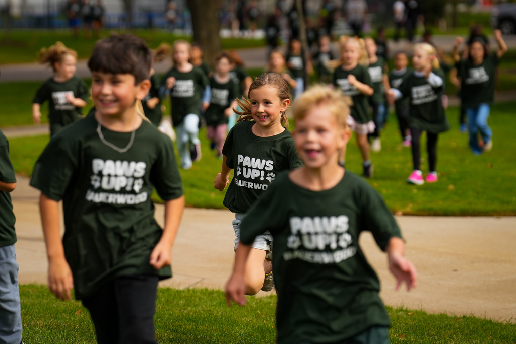 A group of children in green shirts run on grass. The shirts read "PAWS UP".