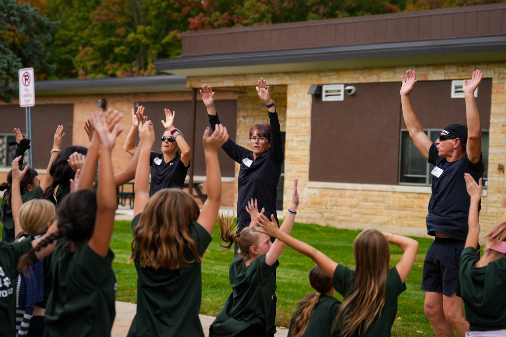 Group of people in green shirts raise hands in unison in front of a building.
