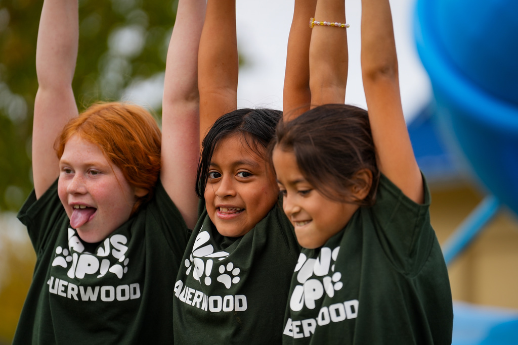 Three children, one with red hair and two with brown, wear matching green shirts. They have their arms raised, hanging on to playground equipment.