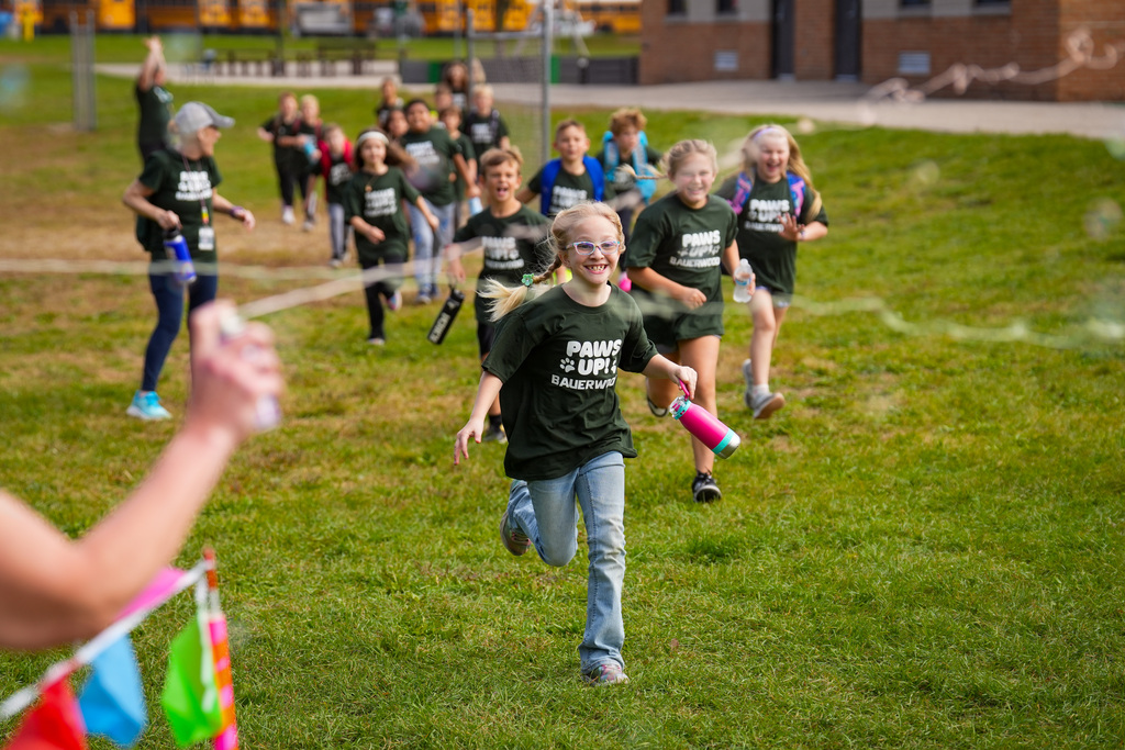 Children in matching T-shirts run on a field with a building in the background.