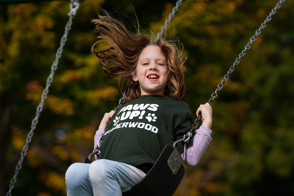 A girl sits on a swing, holding the chains, with hair blowing in the wind, wearing a green t-shirt.