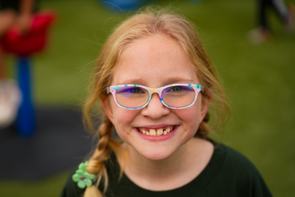 A young girl with braids wearing colorful glasses smiles widely, standing on a green field.
