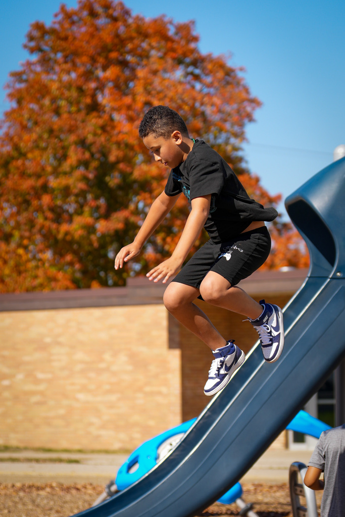 A boy wearing a black T-shirt and shorts jumps on a blue slide at a playground.