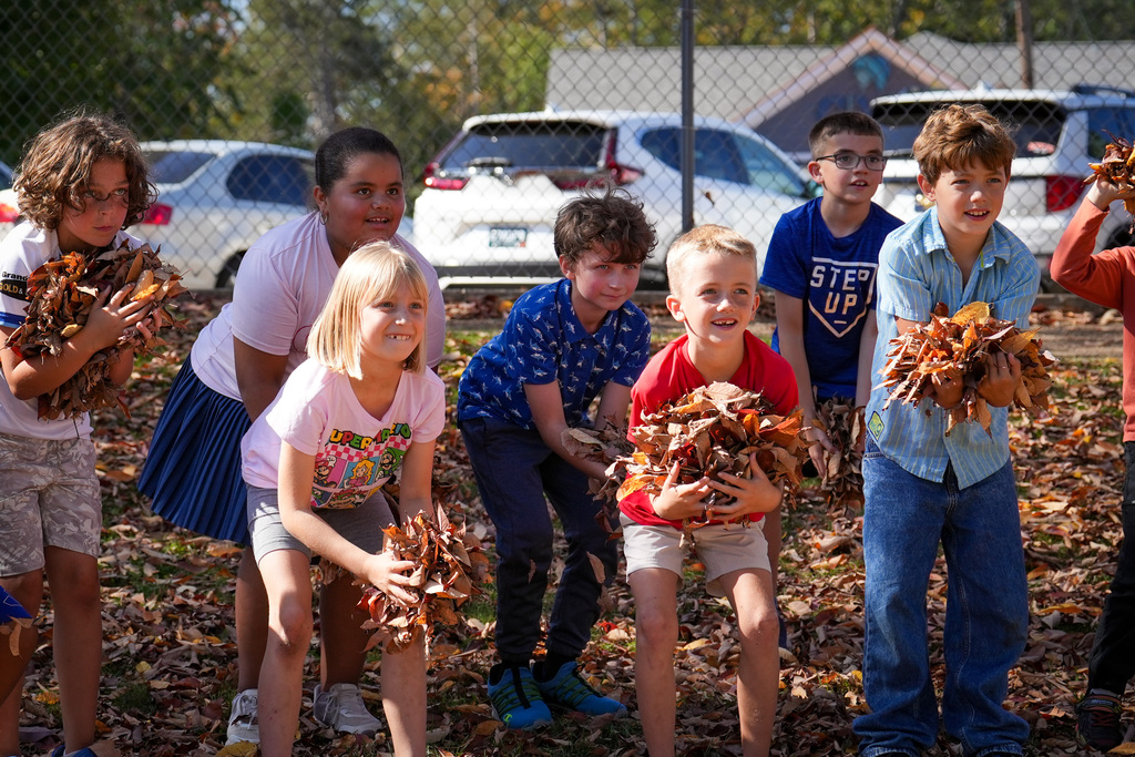 A group of children, some holding leaves, smile as they stand among fallen leaves on a playground..