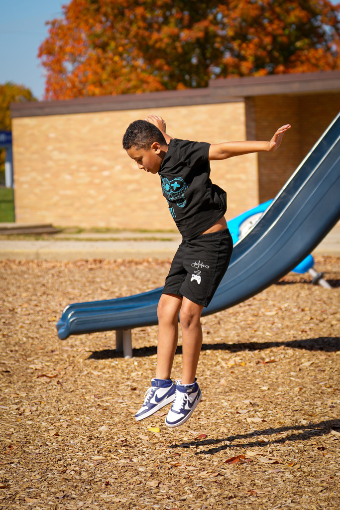 Child jumping from slide on playground, with leaves on ground and trees behind building.