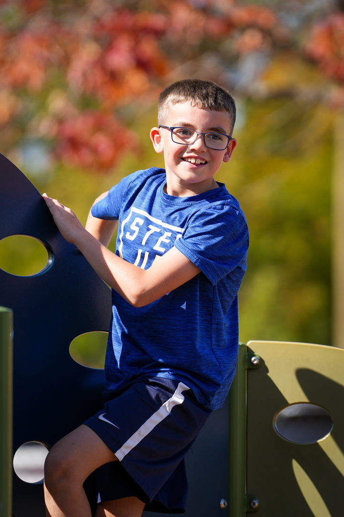 A young boy in a blue shirt and glasses plays on a playground. Green metal structures surround him. Trees with orange leaves are in the background.