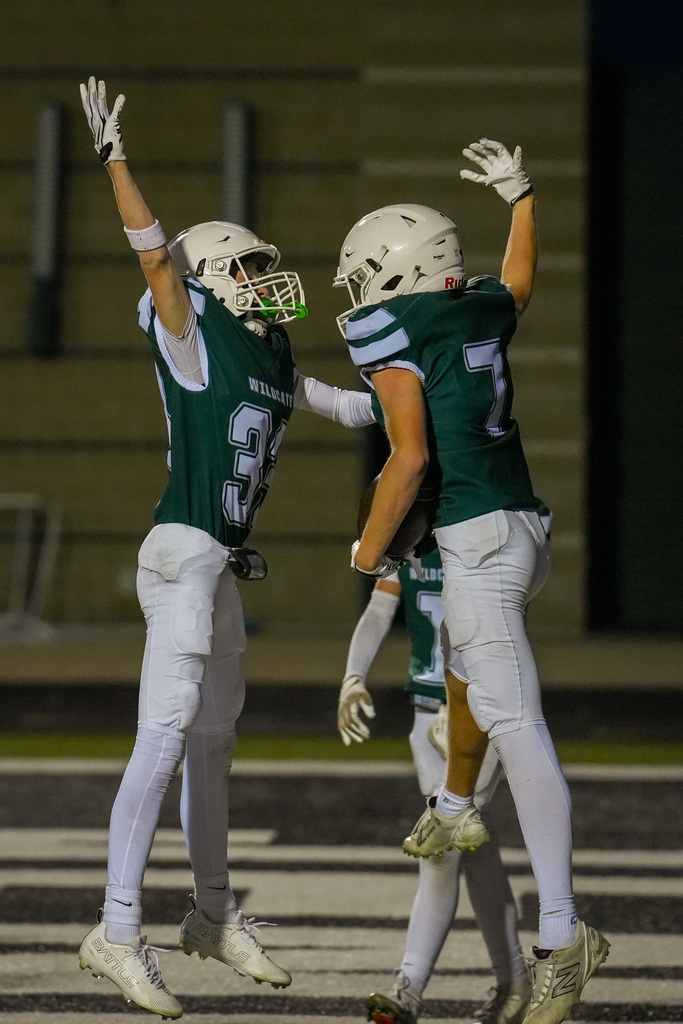 Two football players in green uniforms jump in air on field. One wears helmet and holds hands up.