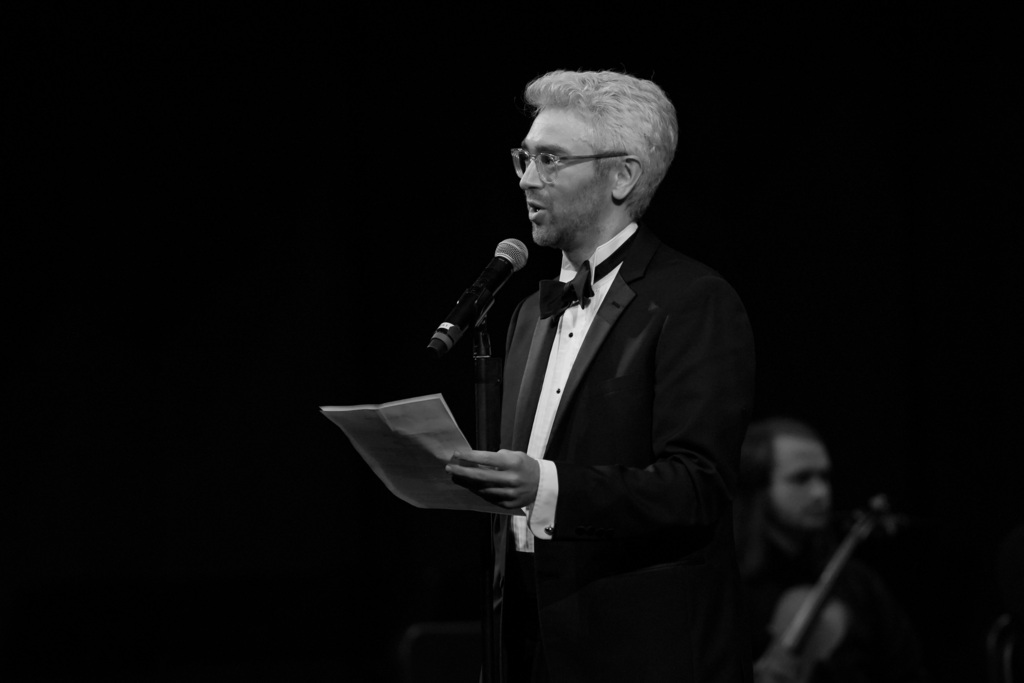 A man with glasses stands at a microphone, holding paper, and speaking. Behind him, another person plays a violin.