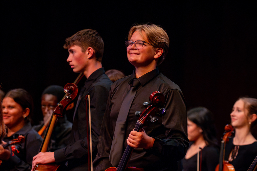 Musicians stand on a stage. A cellist stands in the front. Others stand behind him.