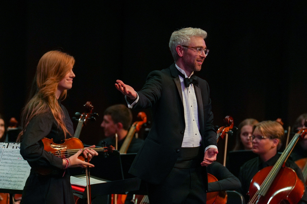 A man in formal attire conducts a woman playing violin while a string ensemble performs in the background.