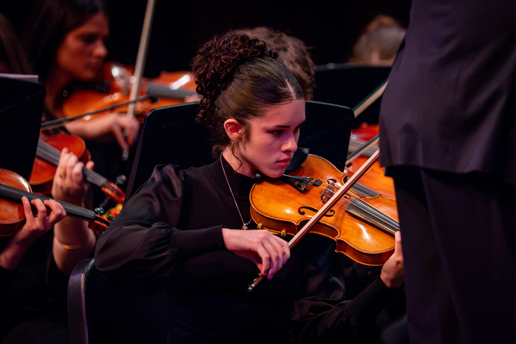 A young violinist in black sits among others playing instruments on stage. They hold the violin under the chin.