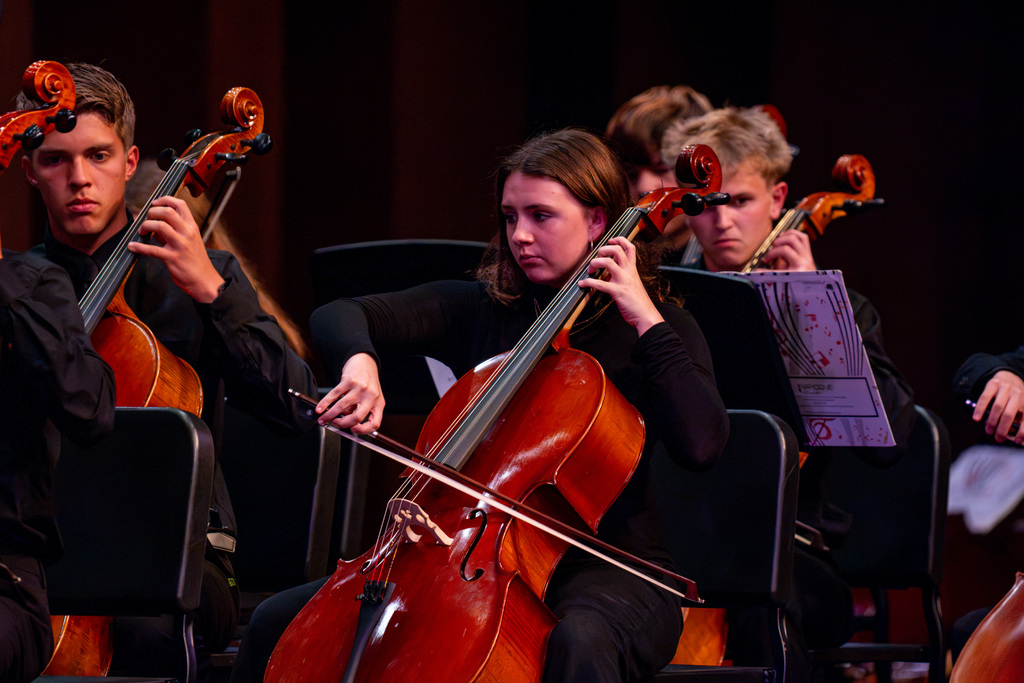 Four people seated, playing cellos, with focus on the woman holding a bow in front of her.