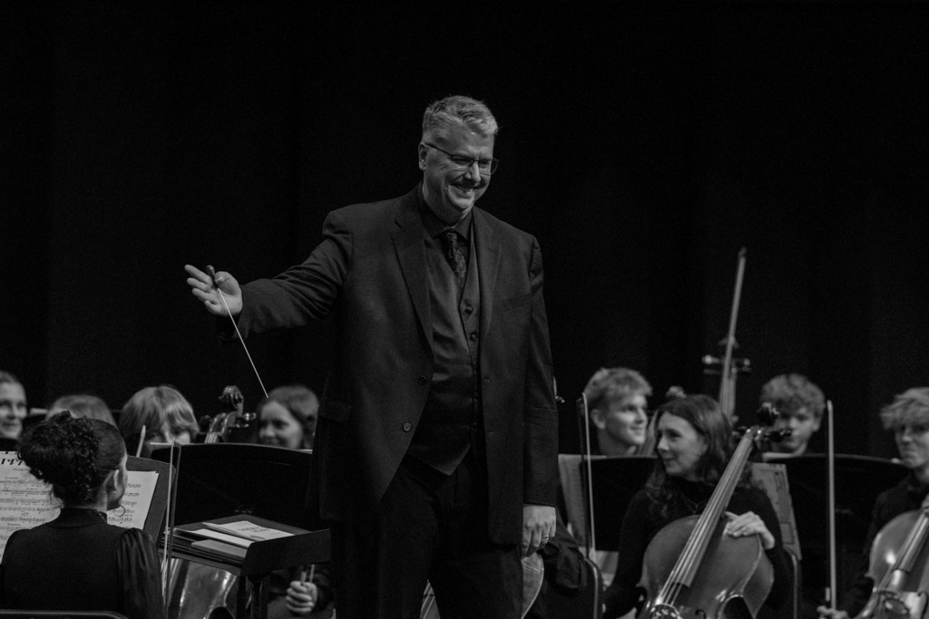A black-and-white photo of a man with a baton standing in front of an orchestra with cellists and violinists.