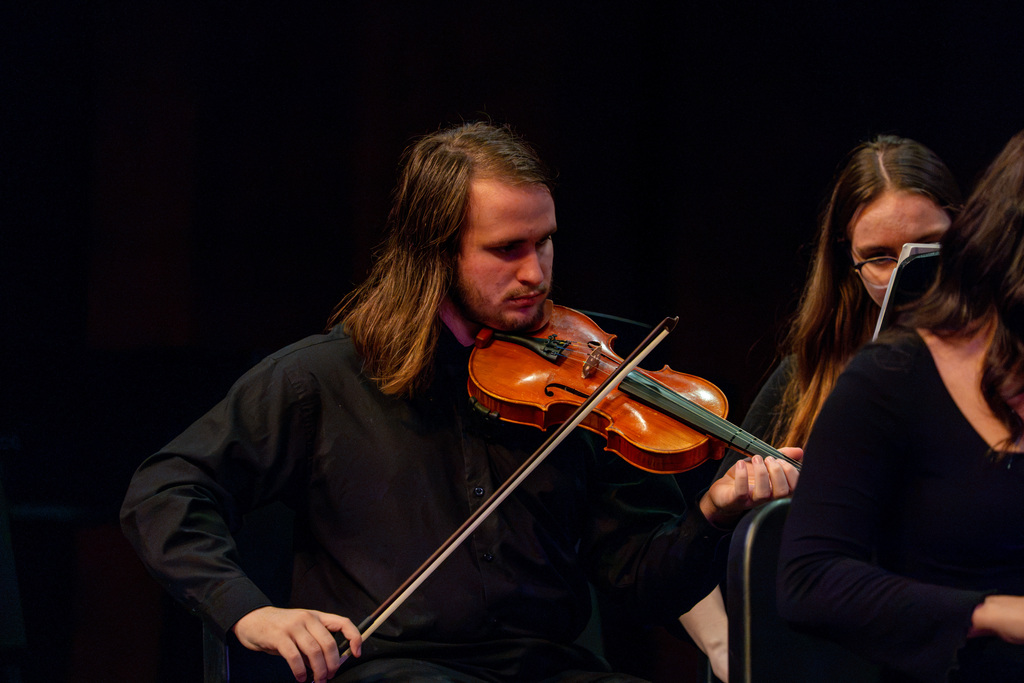 A violinist in black, with long hair, plays in a concert. A woman in black holds a music sheet.