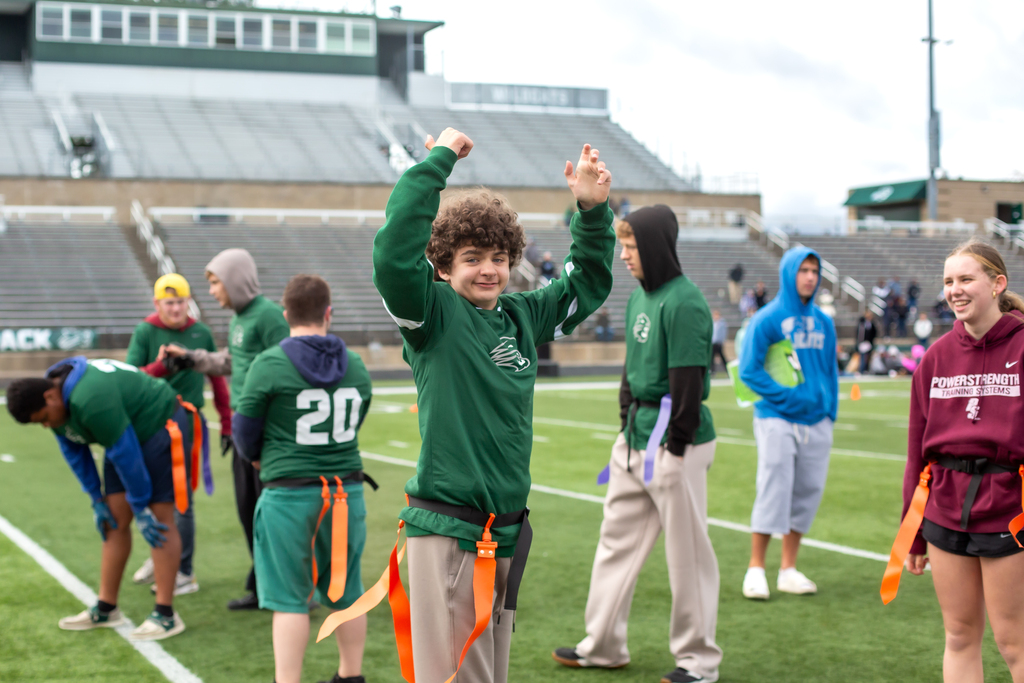 A group of children in green shirts and shorts lined up on a football field. One child is jumping with arms raised.