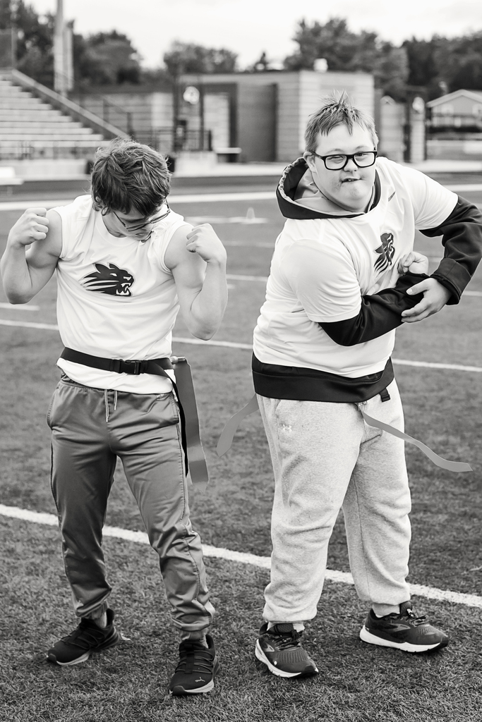 Two boys in sports attire stand on a field. One is flexing. Both wear glasses.