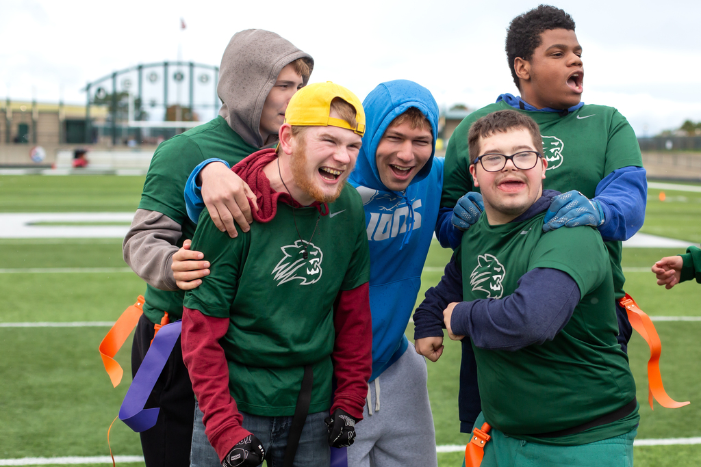 Students in green jerseys and blue hoodies standing on a field with smiles and wide-angle view of a stadium.
