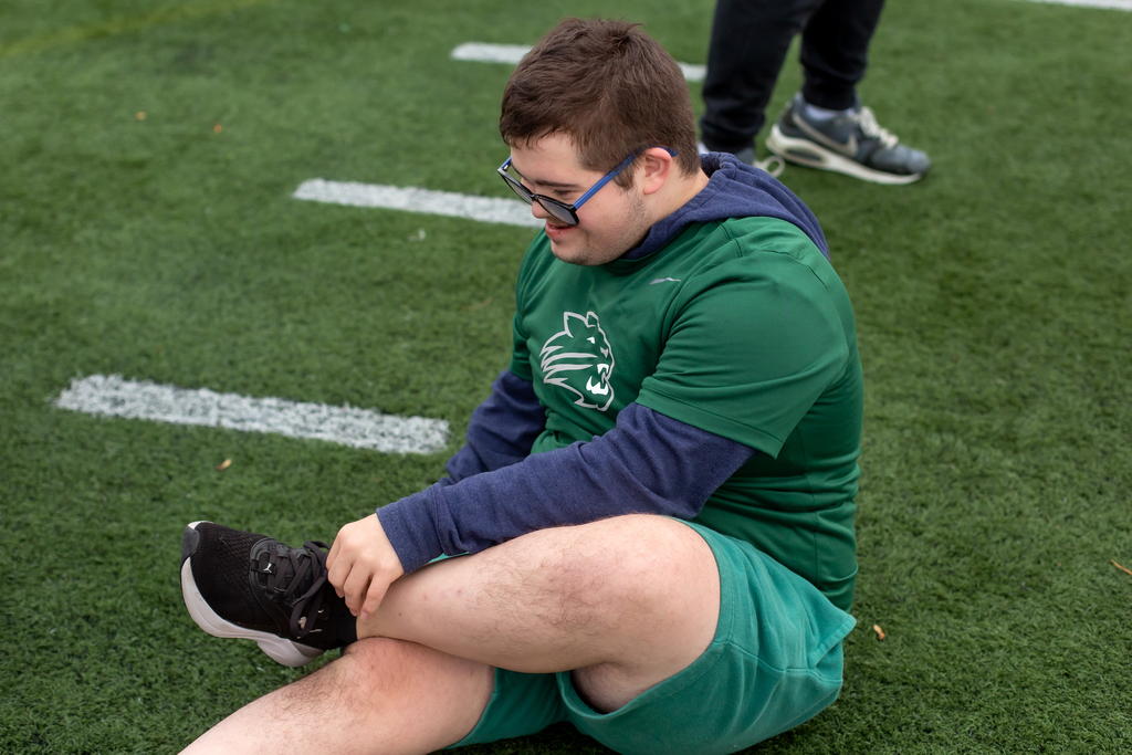 Person in green shirt and shorts sits on grass, adjusts shoe, with white lines on ground.