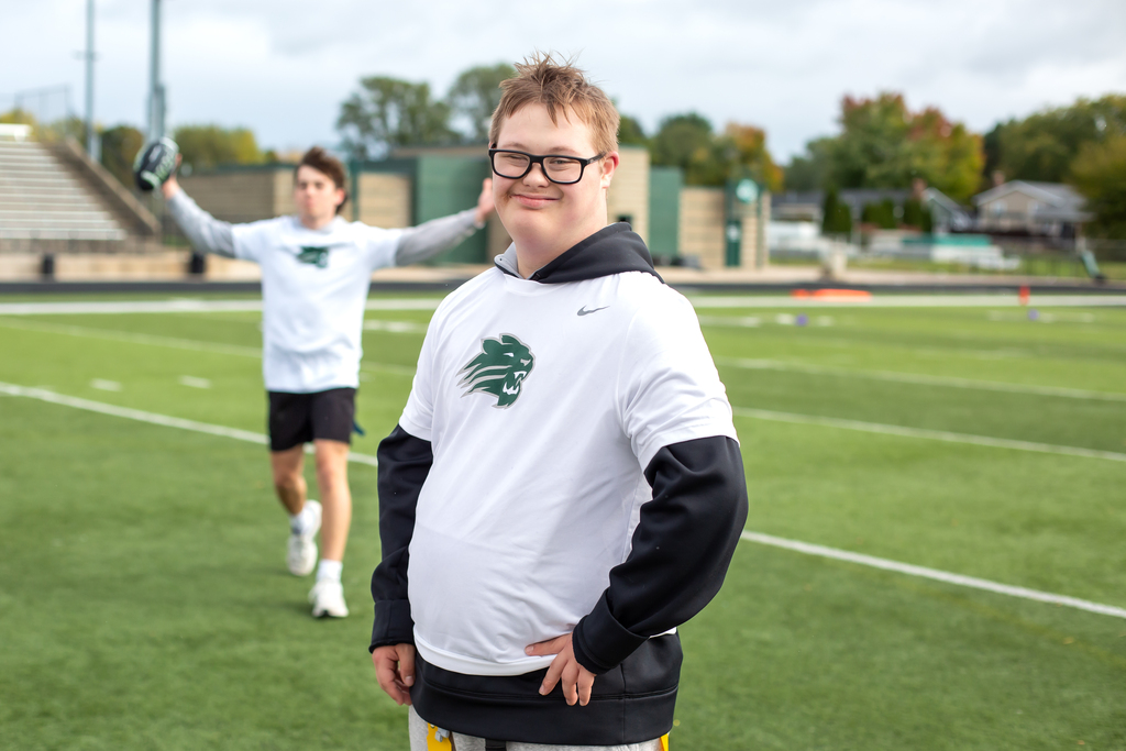 Two individuals on a football field, one standing with hands on hips, another running with a ball.