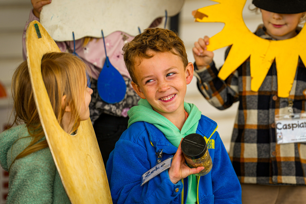 Four kids outside, one holding a jar and smiling, another holding a sun cutout, and two holding large cutouts.