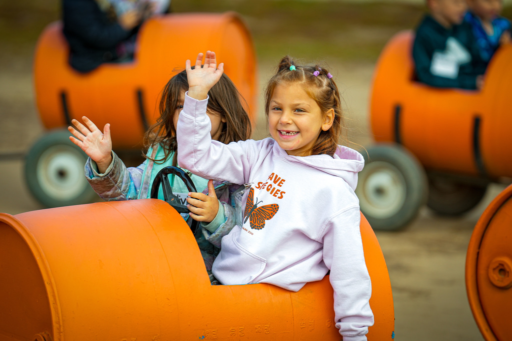 Two children sitting in orange barrel-shaped rides, waving. One child wears a white hoodie.