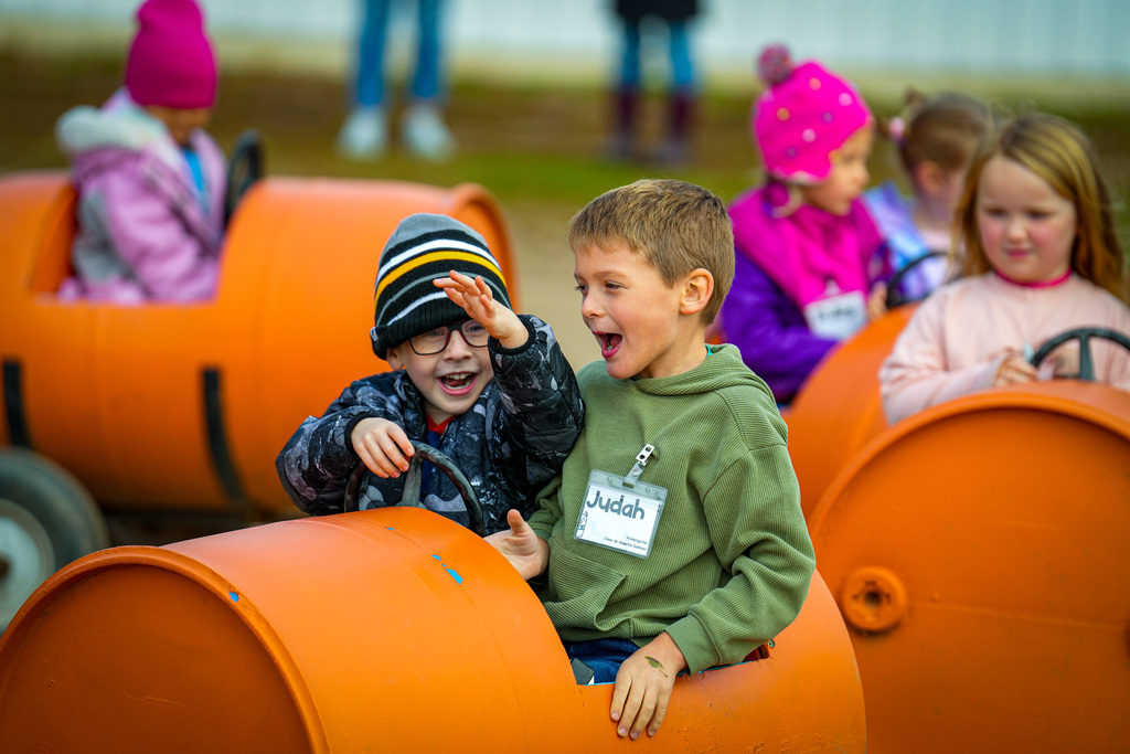 Several young children in winter clothing riding orange barrel-shaped ride with steering wheels.