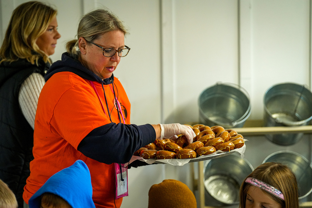 A woman in orange holds a tray of donuts as a group of children looks on.