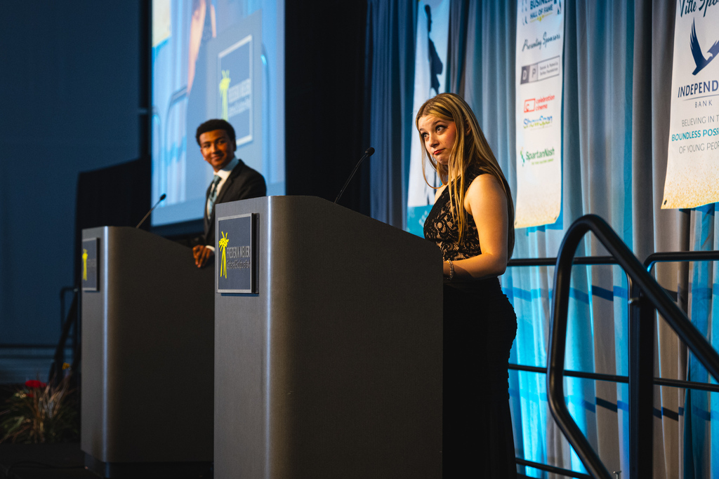 Two students stand at podiums, one with a microphone, while a projector screen displays information in the background.