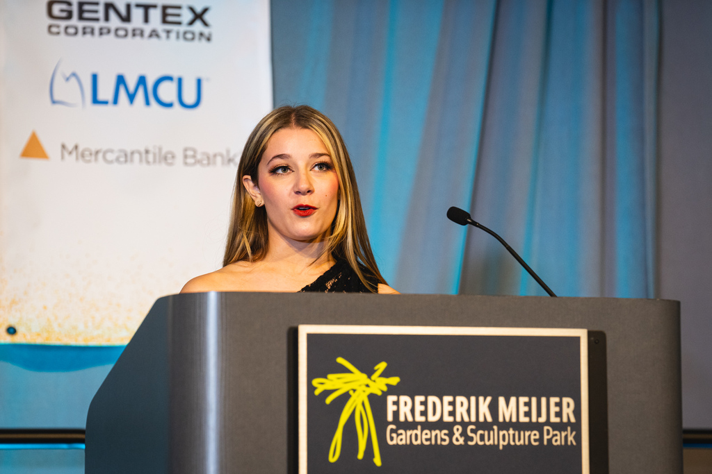 A woman speaking into a microphone at a podium with a sign reading 'Frederik Meijer Gardens & Sculpture Park'.