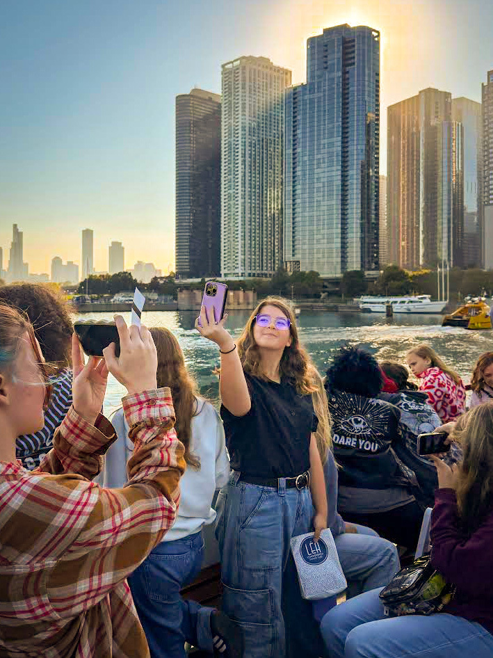Students on a boat by the water, one person wearing glasses and taking a photo.