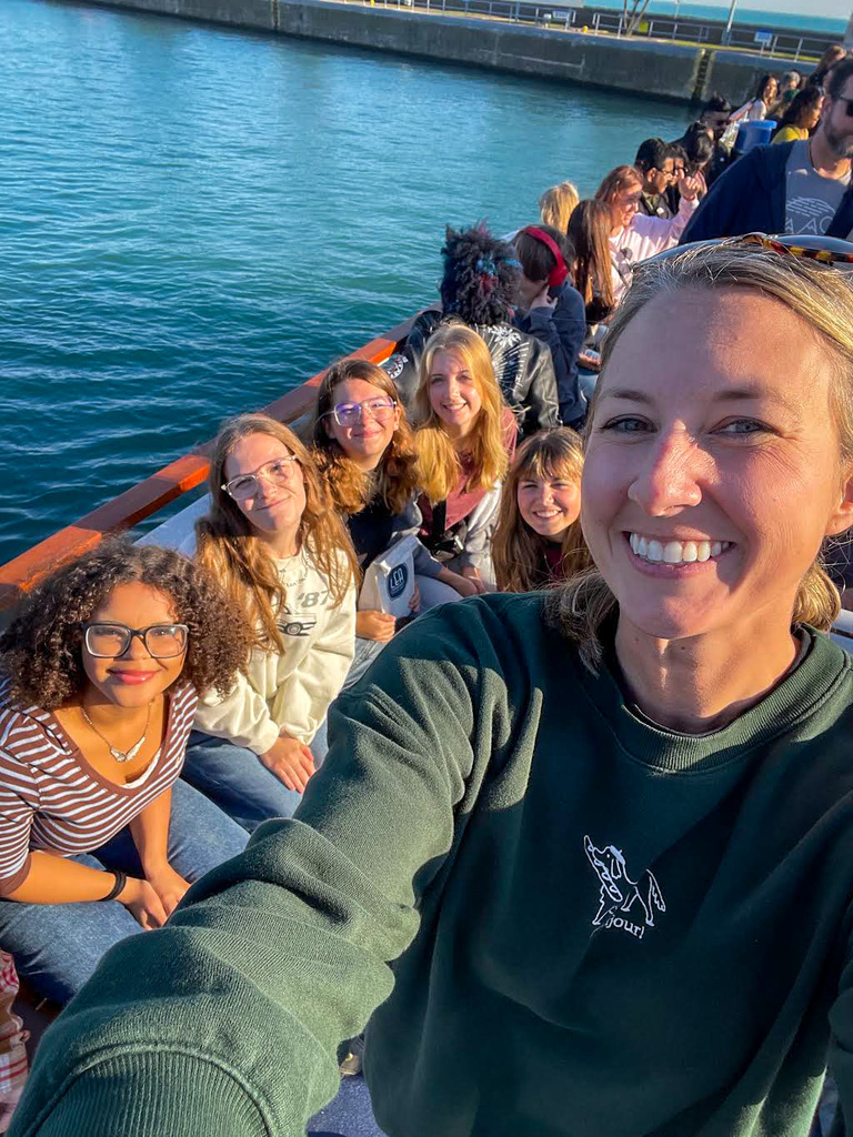 Students on boat on water, staff in green shirt smiles at camera, several students seated behind.