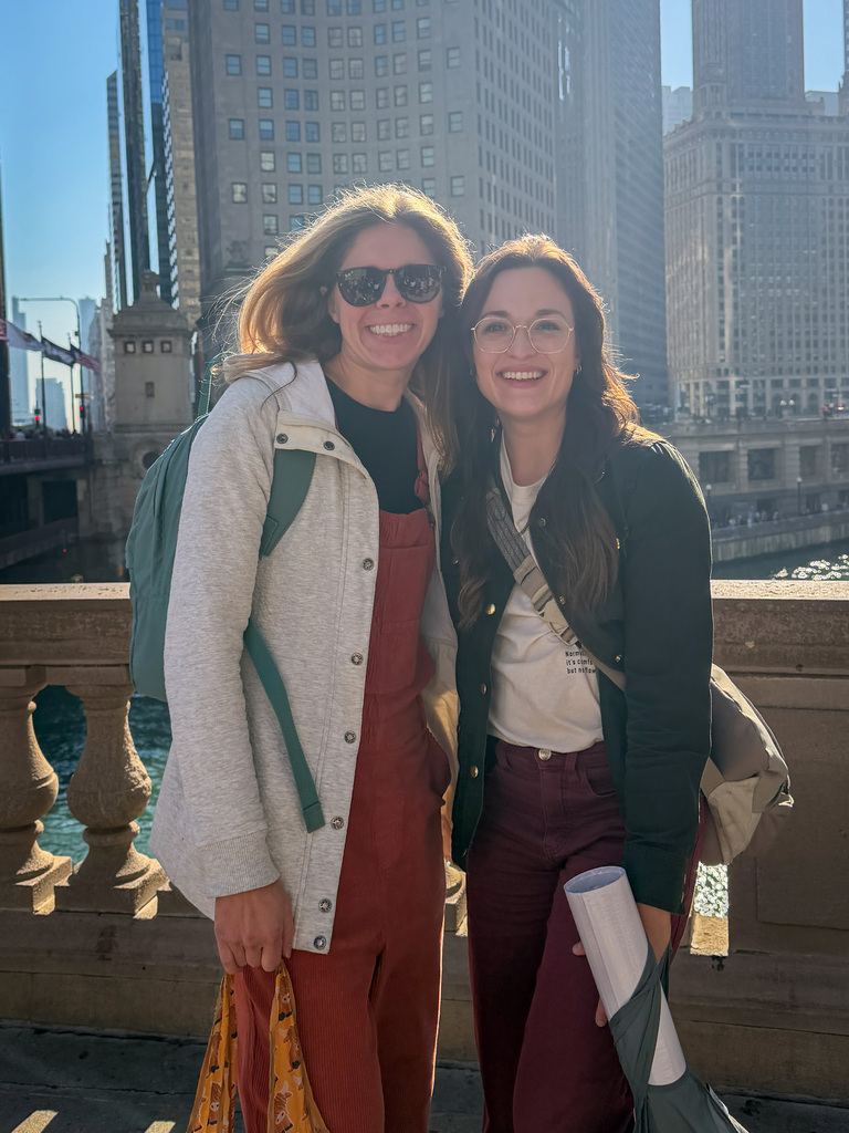 Two staff in casual clothing stand on a bridge with a river and tall buildings in background.