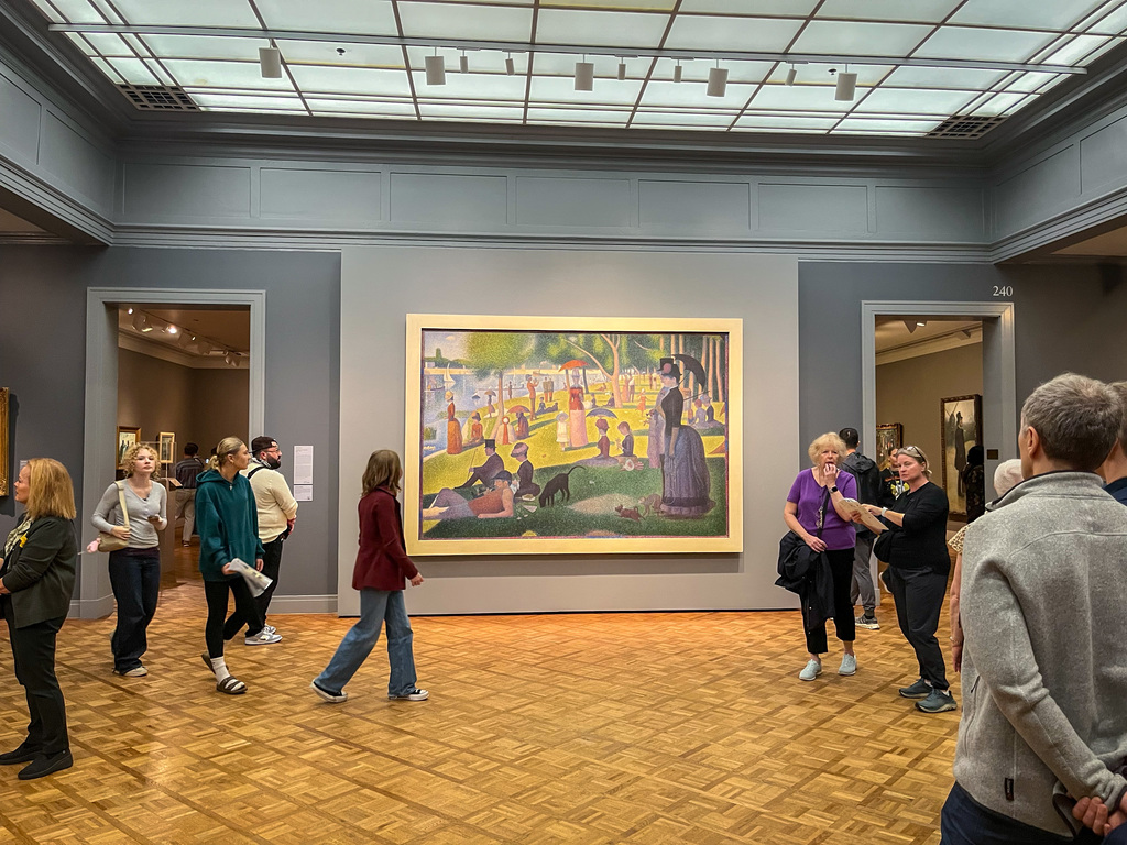 People standing and walking in a museum room with light wooden floors, grey walls, and a large framed painting on one wall.
